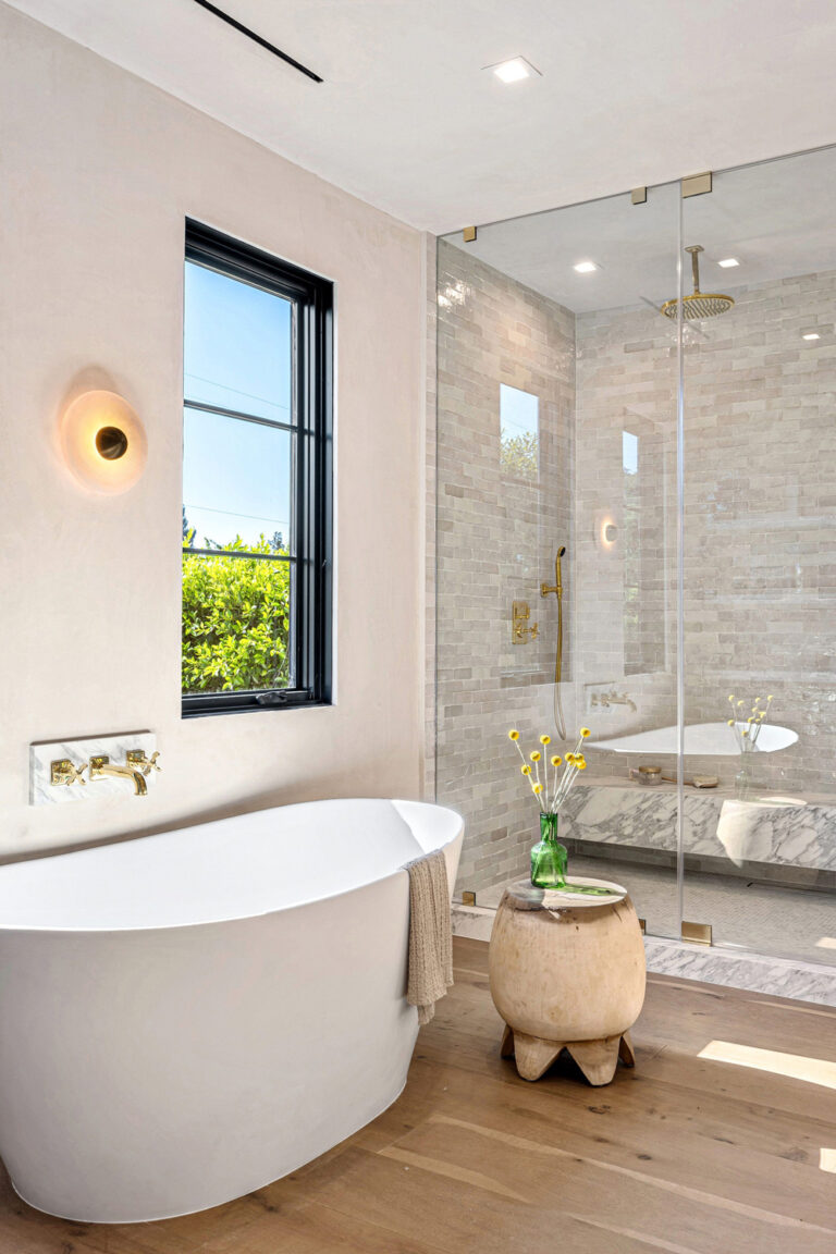 A spa-like bathroom design featuring a white freestanding soaking tub, a large walk-in shower with gray mosaic tiles and gold fixtures, and warm wood flooring.