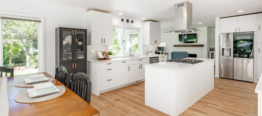 A spacious kitchen and dining area featuring white perimeter cabinets, a contrasting black hutch, a large white island, light wood-style flooring, and views of the backyard through large windows.