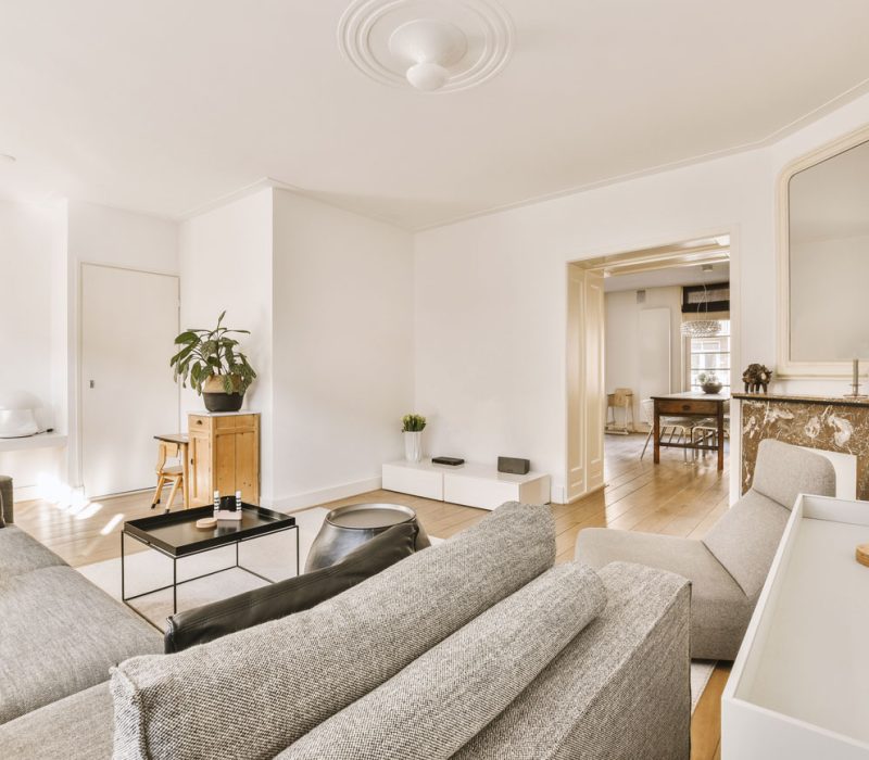 An airy living room furnished with a gray sectional sofa, minimalist side tables, and a marble-topped console, featuring an open doorway that leads to a dining area in the background.