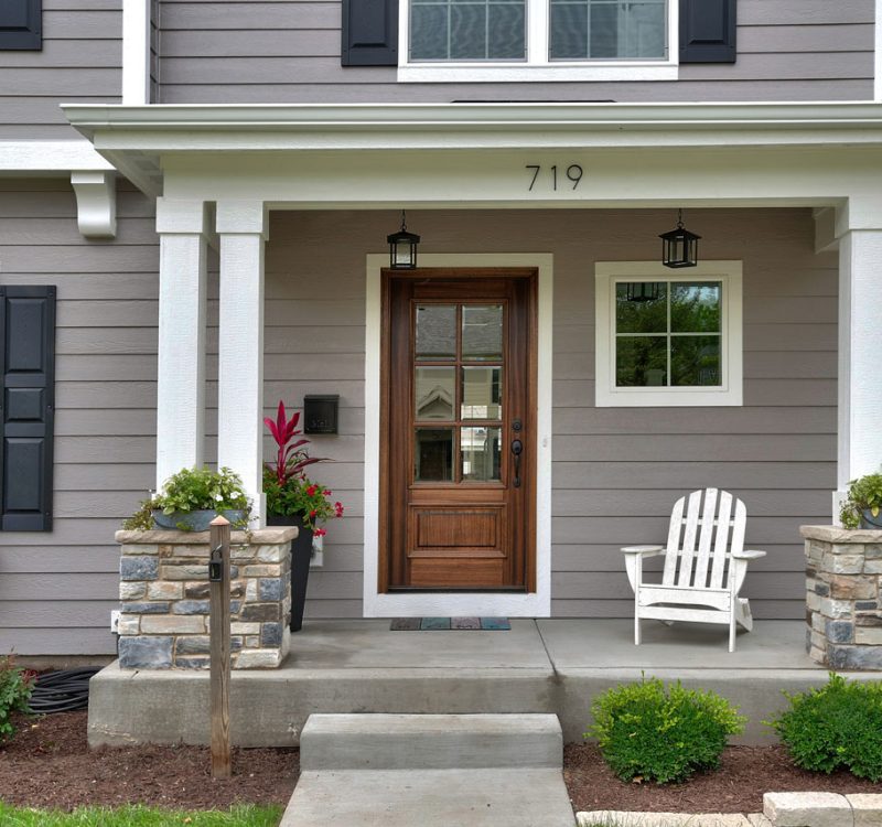 A charming home exterior showcasing a covered front porch with white columns on stacked stone pillars, a custom wood front door, and professional landscaping.